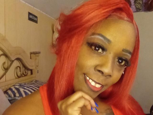 JuiceTiax with bright red hair, dark skin, smiling with blue nails, wearing red, has long eyelashes, and a small nose piercing. Her tits are partially visible.