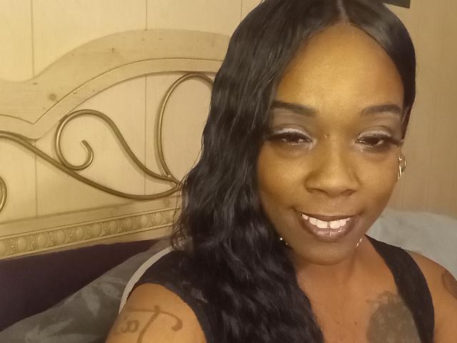JuiceTiax with dark brown skin, black wavy hair, shiny silver eyeshadow, smiling with white teeth, wearing black sleeveless top, showing tattoo on right arm, sitting on bed.