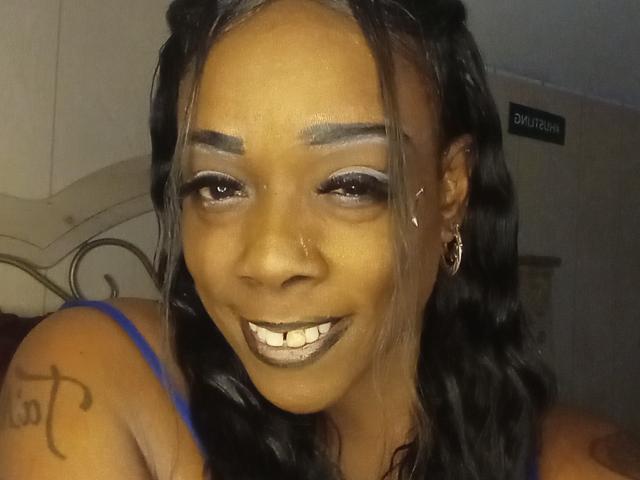JuiceTiax with dark skin, black wavy hair, glossy silver eye makeup, grey lipstick, blue strap dress, nose ring, tattoo on right shoulder, smiling, visible semen on face.