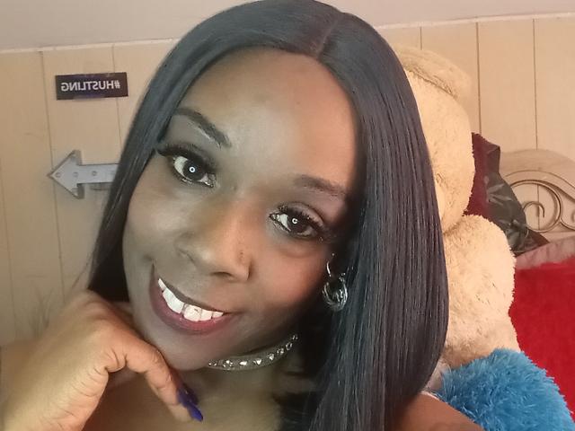 JuiceTiax has dark skin, long black hair, and a cute smile showing white teeth. She's wearing a choker, hoop earrings, and blue nails, with a white stuffed bear behind.