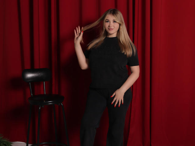 SarahStar staat in zwarte kleding, met haar hand in haar haar, rode nagels, naast een zwarte kruk, met een rode gordijn als achtergrond, lachend, blond haar, middelgrote borsten, slank, schattig..