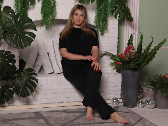 SarahStar, barefoot, black outfit, sitting on gray rug, hands on knees, long blonde hair, red nails, white brick fireplace, green plants, pink flowers, silver balls in background.