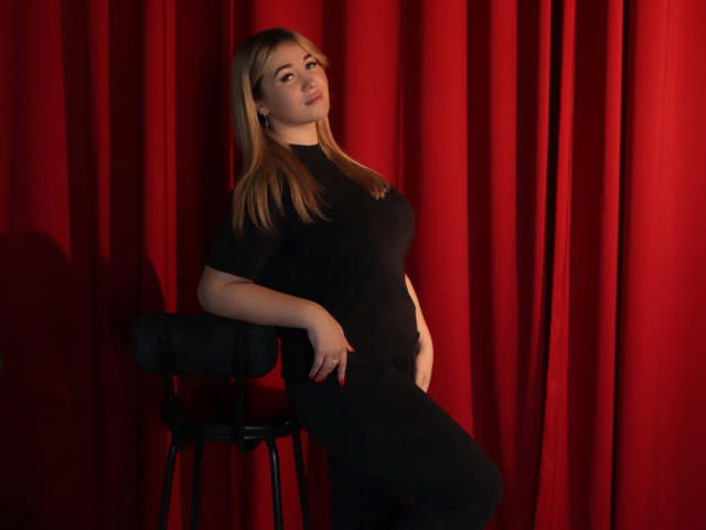 SarahStar mit blonden Haaren, einem schwarzen T-Shirt und einer schwarzen Hose, die an einem Hocker lehnt, rote Vorhänge, ihre Hand auf ihrem Bauch, ihr Hintern leicht angehoben, blickt in die Kamera..