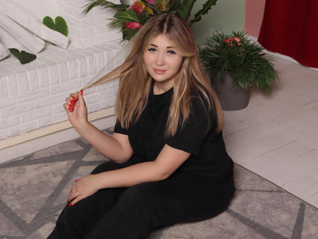 SarahStar with long blonde hair, black dress, and red nails, sitting on a rug, holding her hair with a slight smile, her ass barely visible under her dress.