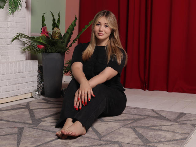 SarahStar, avec ses cheveux blonds, ses vêtements noirs, ses ongles rouges, assise sur un tapis, pieds nus, souriante, devant un mur en briques blanches, avec un rideau rouge, une plante verte et un sol blanc..