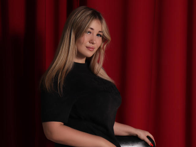 SarahStar with blonde hair, black top, and subtle smile, sitting against red curtains, showing her tits and ass, looking cute with a slight smirk.