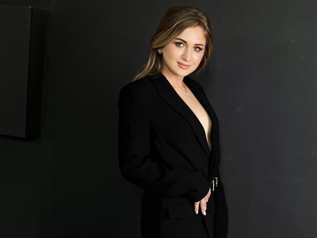 BeataAngel with long brown hair, wearing a black blazer, exposing her tits, standing against a dark background, hands in pockets, smiling subtly, looking at the camera.
