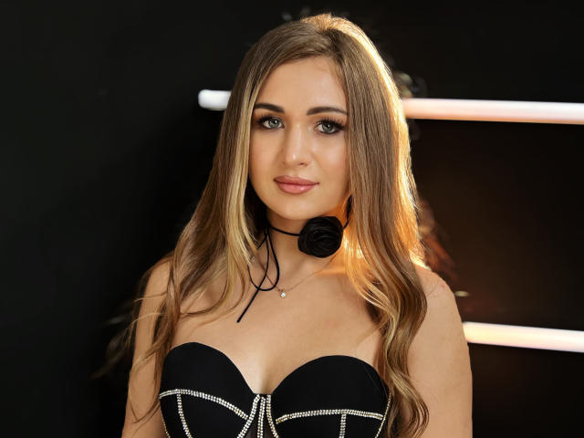 BeataAngel, avec de longs cheveux blonds, portant une robe noire sans bretelles avec des accents argentés, et un collier micro noir, souriant à la caméra, montrant ses seins..