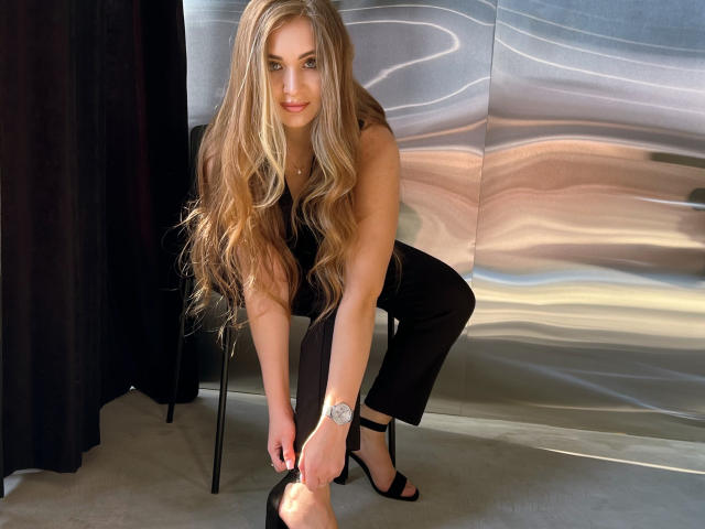 BeataAngel with long blonde hair, black sleeveless top, and black pants, bending to tie black high heels, showing her slim, sexy ass, and a silver watch on her wrist.