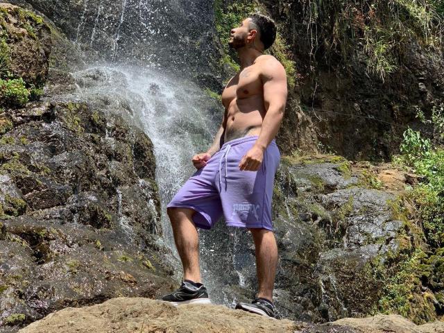 Shirtless MarkBilly69 with a muscular build, small nipples, purple shorts, and black sneakers standing in front of a waterfall, water splashing around his legs.