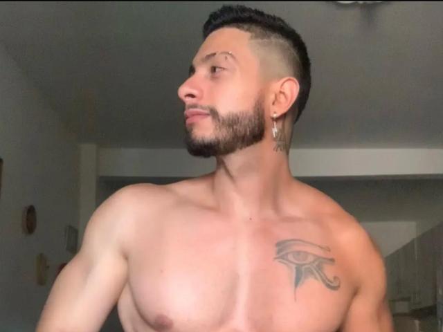 Shirtless MarkBilly69 with a tattoo of an eye on his right chest, short black hair, beard, ear gauges, and side profile, looking to the left.