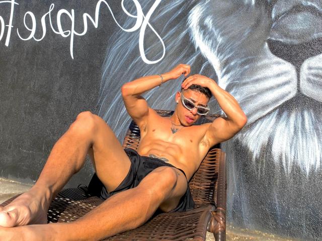 Pjao, muscular, tan-skinned, wearing black shorts and sunglasses, lounging on a wicker chair, hands in hair, large ass, tattoo on chest, graffiti with lion face in background.
