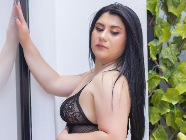 AbbyRed with black lace lingerie, touching a white wall, has long black hair, full tits, and a serious expression, green leafy plant in background.
