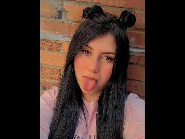 AbbyRed has long black hair in two buns, brown eyes, a nose piercing, and sticks her tongue out. She's wearing a pink shirt against a brick wall.