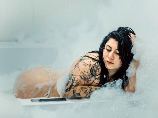 AbbyRed mit schwarzen Haaren, Tätowierungen am Arm und Rücken, liegt nackt in einer weißen Badewanne voller Schaum, mit einem ruhigen Gesichtsausdruck, wobei ihr Hintern teilweise von Schaum bedeckt ist..