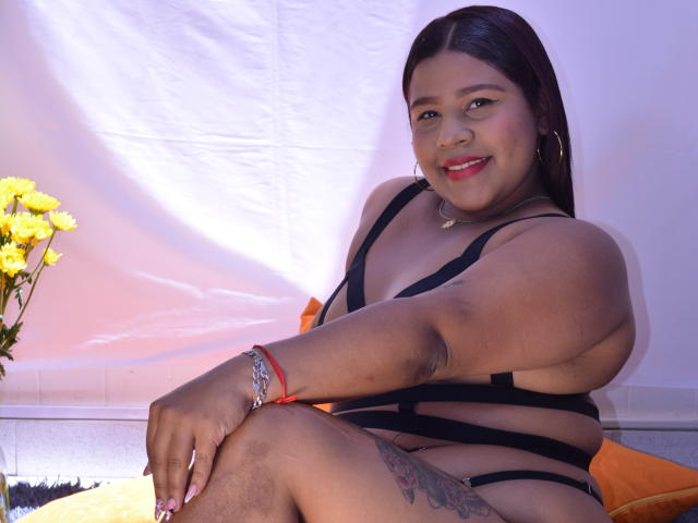 PamelaMckein with dark skin, red lipstick, black strappy lingerie, smiling, sitting, visible tattoo on thigh, yellow flowers, orange pillow, red bracelet, and purple lighting.