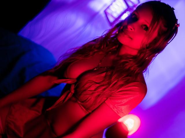 KeiliSexy with red-lit skin, long wavy hair, tied-up shirt exposing her tits, looking seductively at the camera, standing in a purple-blue light, with a glowing lamp.