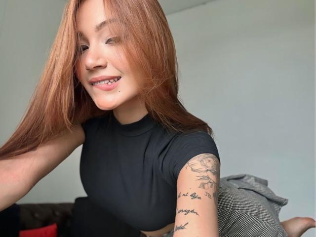MadelineEgan has long red hair, black crop top, tattooed arm with rose and text, smiling with small nose piercing, sitting on gray pillow. Her tits are not visible.
