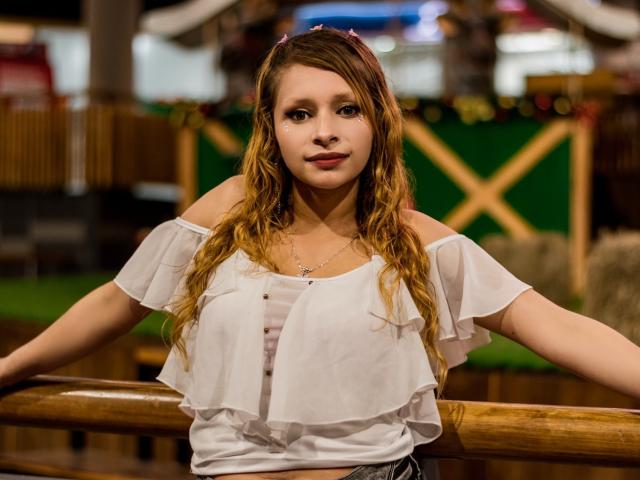 KeiliSexy with wavy blonde hair, off-shoulder white ruffle top, small tits, and a necklace, leaning on a wooden railing, looking directly at the camera.