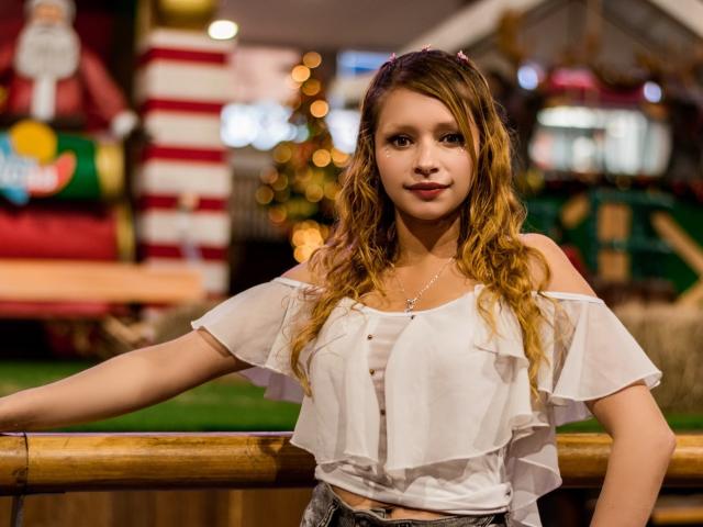 KeiliSexy, with wavy brown hair, wears a white off-shoulder top, showing small tits, and stands against a bamboo railing, looking cute with a slight smile.