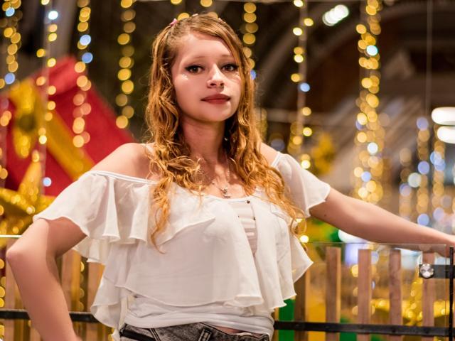 KeiliSexy with wavy blonde hair, wearing a white off-shoulder top, hand on railing, looking forward, soft smile, bokeh lights in background, slight blush on cheeks, subtle makeup.