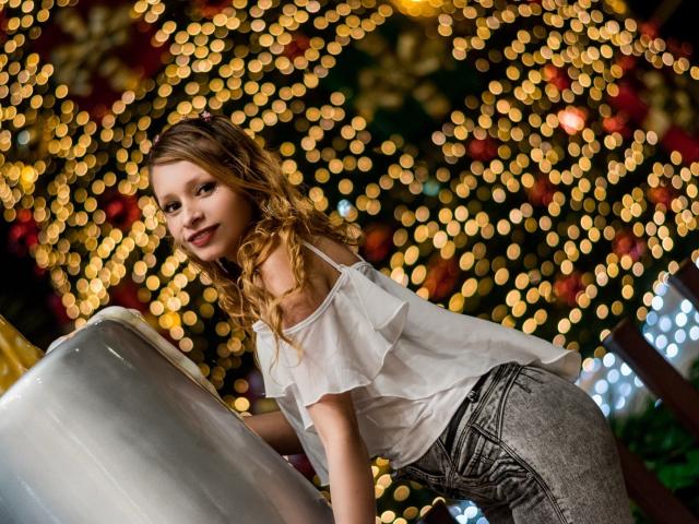 KeiliSexy with curly blonde hair, white ruffled top, and gray pants, leaning on a silver metal cylinder, smiling, bokeh lights in background.