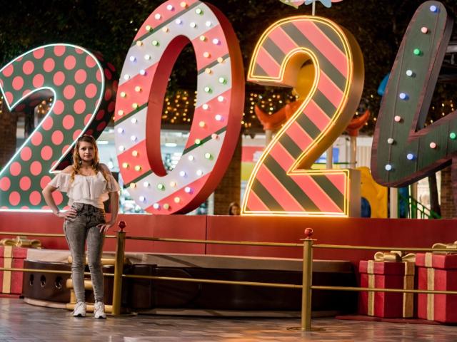 KeiliSexy stands in front of giant, colorful "2022" letters. She has long brown hair, a white blouse, gray pants, and yellow shoes. Gift boxes are on the ground.