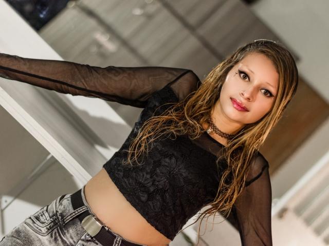 KeiliSexy with wet blonde hair, black lace crop top, sheer sleeves, and gray jeans, wearing a choker, leaning on a white surface, smiling, with red lipstick and dark eyeliner.