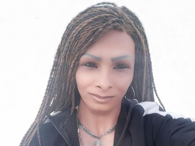 SiliconBoobs tem cabelo trançado marrom, sobrancelhas escuras e está vestindo uma jaqueta preta com listras brancas. Ela está sorrindo, usando um colar de prata e brincos de argola..