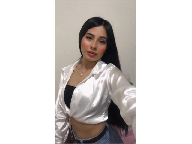 ValeryBell, con i suoi lunghi capelli neri, indossa una giacca di raso bianco lucida, un reggiseno nero e jeans blu. Ha un viso carino, un seno piccolo e un sorriso sicuro..