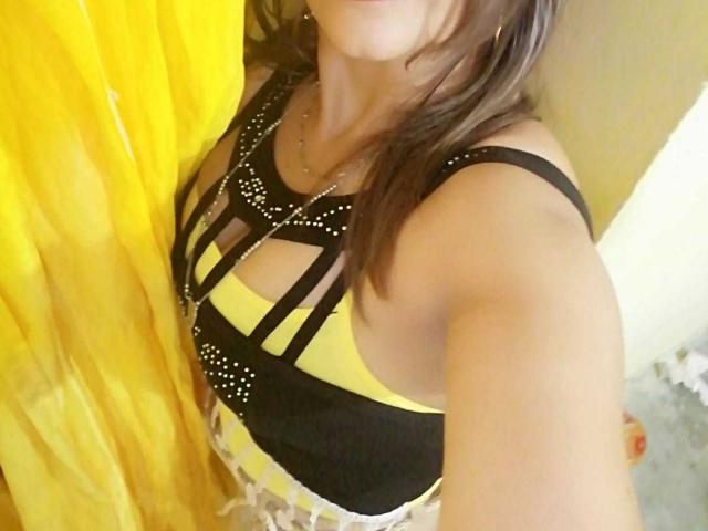 EstrellitaSexy con un seno di media grandezza, indossa un abito nero e giallo con spalline sottili, c'è una tenda gialla a sinistra, ha i capelli castani e una collana d'oro, e sta scattando un selfie..