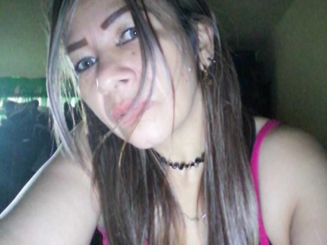 EstrellitaSexy, avec ses lèvres pulpeuses, ses cheveux bruns, son haut rose, son collier noir et ses boucles d'oreilles, regardant vers le haut avec les lèvres légèrement entrouvertes, et un petit anneau de nez, dans une pièce faiblement éclairée..