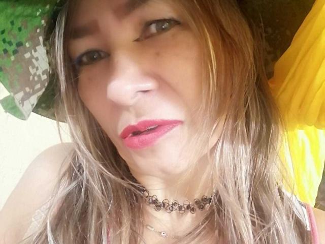 EstrellitaSexy con cabello rubio despeinado, un gorro de camuflaje verde, lápiz labial rosa, un collar negro y una cortina amarilla de fondo, con una expresión ligeramente molesta. Se puede ver el contorno de los senos debajo de la blusa rosa..