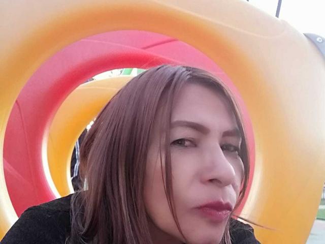 EstrellitaSexy tem cabelo castanho, olhos verdes e está fazendo beicinho com os lábios vermelhos. Ela está em frente a um aro inflável amarelo e vermelho. Ela está vestindo uma blusa preta..