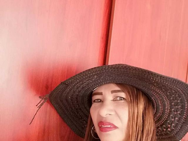 EstrellitaSexy indossa un cappello intrecciato nero, rossetto rosso e orecchini a cerchio, con capelli castani e sopracciglia scure, sullo sfondo di un muro di legno rosso..