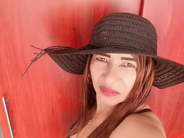 EstrellitaSexy with brown hair, black woven hat, pink lipstick, hoop earrings, and a necklace, showing her face and upper chest. Red background. Boobs partially visible.