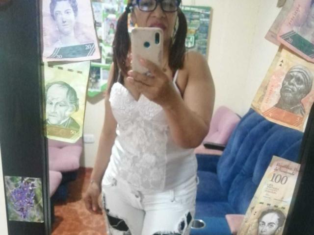 EstrellitaSexy with dark skin, glasses, and pigtails, taking a mirror selfie. She's wearing a white lace tank top and white pants, showing her ass in the mirror.