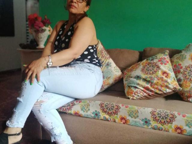 EstrellitaSexy with medium boobs, wearing a black polka dot top and ripped light blue jeans, sitting on a floral-patterned couch, green nails, and a wristwatch.