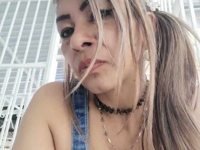 EstrellitaSexy, avec ses couettes blondes, portant une salopette en jean et un collier noir, a une expression boudeuse, un nez visible et de petites boucles d'oreilles, sur une photo de profil légèrement désordonnée..
