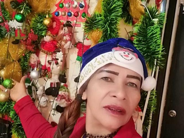 EstrellitaSexy che indossa un cappello da Babbo Natale con la faccia di un pupazzo di neve, rossetto rosso e una camicia rossa, che alza un dito, con decorazioni natalizie tra cui festoni verdi e dorati..