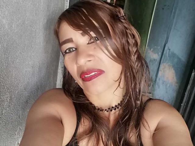 EstrellitaSexy hat feuchtes, braunes Haar, roten Lippenstift, eine schwarze Halskette und blickt in die Kamera, wobei ihre Brüste leicht unter einem schwarzen Oberteil zu sehen sind..