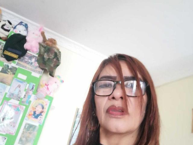 EstrellitaSexy, com cabelo castanho, óculos e lábios carnudos, tem um nariz pequeno e está olhando para a câmera com uma leve carranca, em um quarto claro..