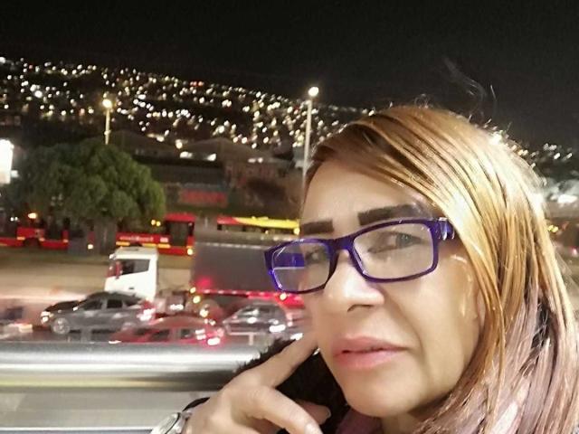 EstrellitaSexy, avec des lunettes bleues, des cheveux châtains clairs et des lèvres roses, tenant son téléphone, dans un environnement urbain nocturne avec des lumières de voitures floues en arrière-plan..