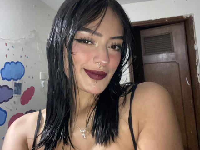 MiraCataleyax has black hair, dark eyeliner, maroon lipstick, nose ring, lip ring, and a necklace. She's wearing a black spaghetti strap top, showing her tits.