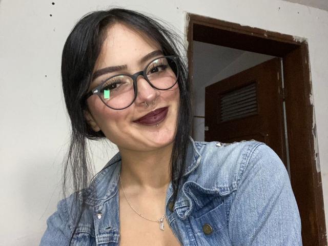 MiraCataleyax with black hair, glasses, nose ring, and dark lipstick, wearing a blue denim jacket, smiling at the camera, reflection in glasses, casual selfie.