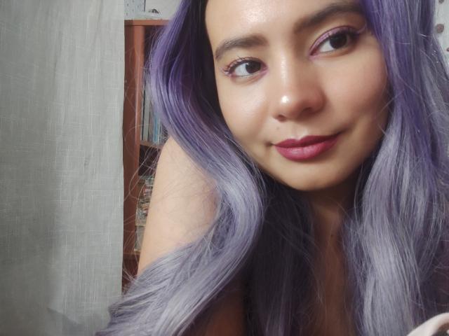 SuzzyV with purple hair, pink eyelids, and pink lipstick, smiling at the camera, showing off her cute face and small, round nose, with her tits partially visible.