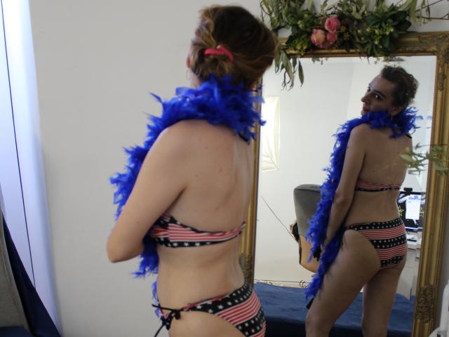 EvelynAngel in a patriotic bikini, blue feather boa, standing in front of a mirror, reflecting her ass and back, with her head turned, smiling at the camera.