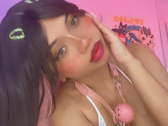 LiliMays with dark hair, pink hair clips, red lipstick, pink collar with ball, hand on cheek, green eyes, white top, pink and purple background. Boobs visible.