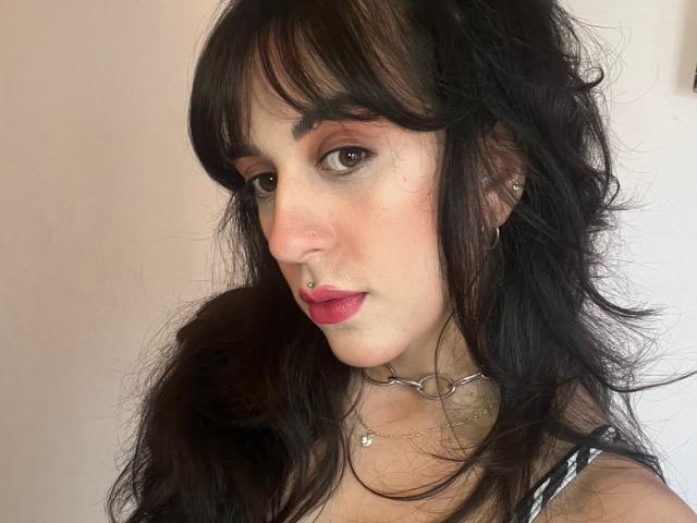 LunaStella has messy black hair, brown eyes, pink lipstick, nose ring, and a silver necklace with heart pendants. She's looking at the camera with a slight smile.
