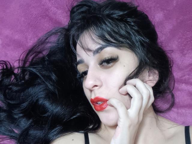 LunaStella has black, wavy hair, pale skin, red lipstick, and her hand near her mouth. She's looking down with long eyelashes, on a pink, fuzzy surface.